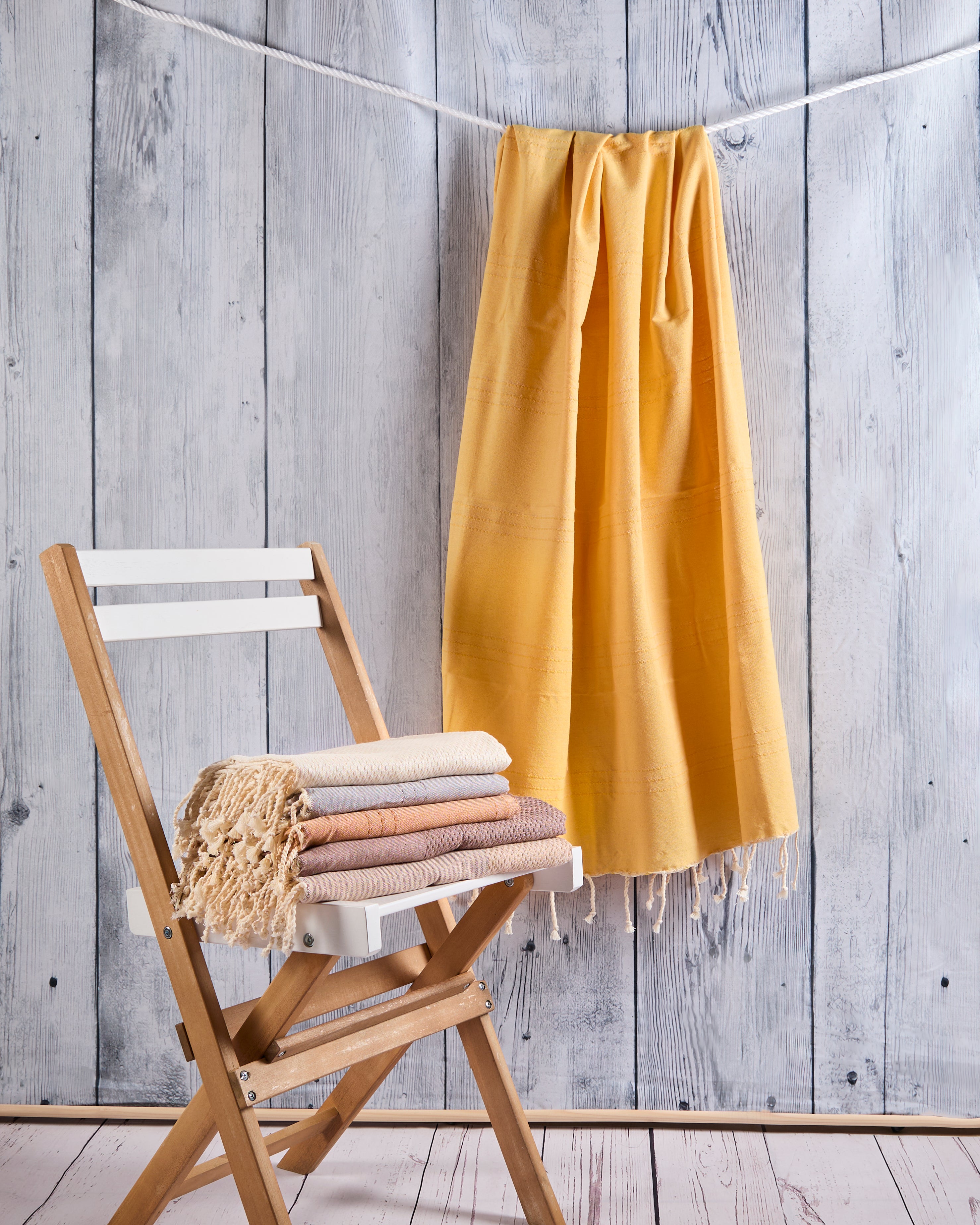 Fouta tunisienne artisanale jaune moutarde suspendue, serviettes roulées sur une chaise en bois, fond en bois naturel.
